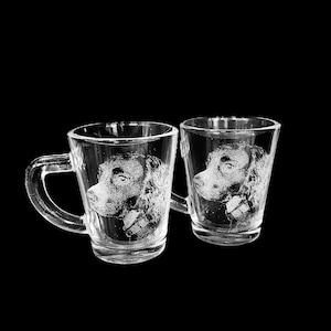 May include: Two clear glass mugs featuring an etched portrait of a dog on the front surface. Each mug has a sturdy handle and a tapered cylindrical shape. The detailed white engraving depicts the side profile of a canine with a collar.
