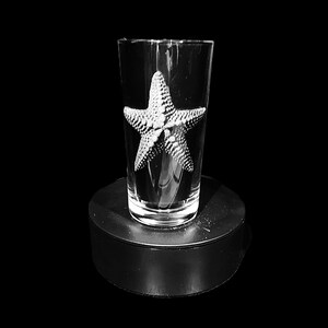 May include: A clear glass tumbler featuring a textured starfish design. The starfish is centrally placed on the glass and appears to be made of a light-coloured material. The glass is on a black circular base.