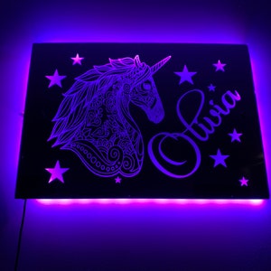 以下が含まれることがあります： 白いユニコーンのデザインと「Olivia」という名前が筆記体で書かれた黒いアクリル製のライトアップサイン。サインは紫色のLEDライトで照らされています。