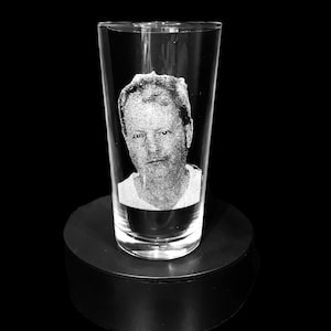 May include: A clear pint glass with a portrait etched into the glass. The portrait is of a person wearing a white shirt. The glass is on a black circular stand against a black background.