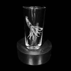 Op de afbeelding: Een helder glazen tumbler met een gedetailleerd geëtst ontwerp van een octopus. Het octopusontwerp is gecentreerd op het glas, met zijn tentakels naar beneden. Het glas staat op een zwarte, ronde standaard tegen een zwarte achtergrond.