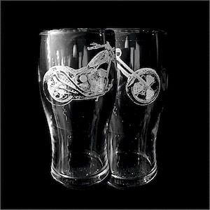 May include: Two clear beer glasses with a motorcycle design etched on the front. The motorcycle is detailed with visible wheels, handlebars, and engine components. The glasses are set against a black background.