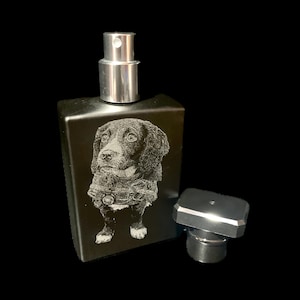 May include: A black rectangular perfume bottle with a silver spray nozzle and a matching cap. The bottle has an engraved image of a dog wearing a jacket. The cap is detached and placed beside the bottle.