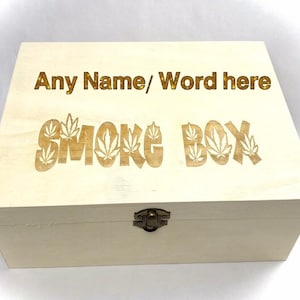 May include: A light-coloured wooden box with the words "Any Name/ Word here" and "SMOKE BOX" engraved on the lid. The text is in a gold colour. The word "SMOKE" has cannabis leaf designs. The box has a metal clasp.