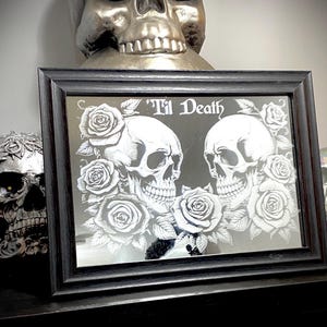 Op de afbeelding: Zwart omrande spiegel met een gotisch ontwerp. De spiegel toont twee schedels omringd door rozen, met de tekst "'Til Death" bovenaan. Het ontwerp is wit op een zwarte achtergrond.