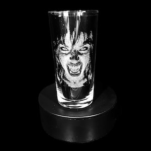 Puede incluir: Un vaso de vidrio transparente con una imagen grabada de una cara gritando. La imagen muestra los ojos, los dientes y el cabello en detalle. El vaso cilíndrico se encuentra sobre una base negra y redonda. El fondo es negro.