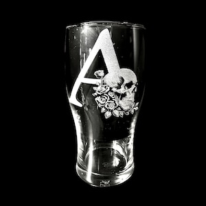 May include: A clear glass with a black background, featuring a decorative design. The design includes a large letter "A", a skull, and roses. The skull and roses are in a detailed, etched style, creating a gothic aesthetic.