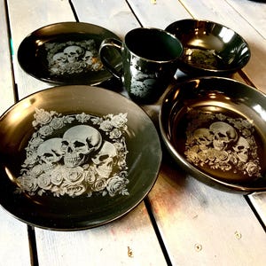 May include: A black ceramic dinnerware set with a skull and rose design. The set includes plates, bowls, and a mug. The skull design is etched in a light colour against the dark background. The set is on a wooden surface.