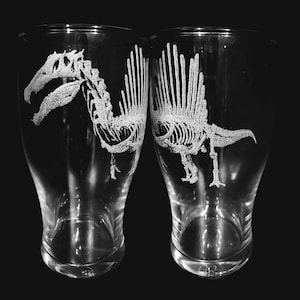 Può includere: Due bicchieri da pinta trasparenti con disegni di scheletri di dinosauri incisi. Gli scheletri bianchi sono dettagliati su uno sfondo nero. I bicchieri sono leggermente curvi e hanno un'ampia apertura.