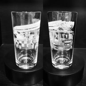 May include: Two clear drinking glasses with etched designs of a truck. The truck design is detailed, showing the front, wheels, and top of the vehicle. The glasses are displayed on a black surface.