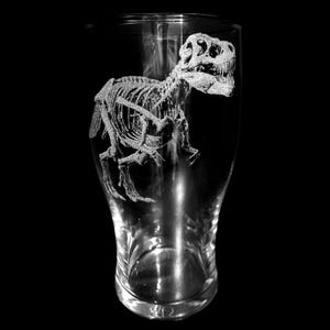 May include: A clear pint glass featuring an etched design of a Tyrannosaurus Rex skeleton. The dinosaur skeleton is shown in profile, with its bones and skull visible. The glass is set against a black background, highlighting the design.