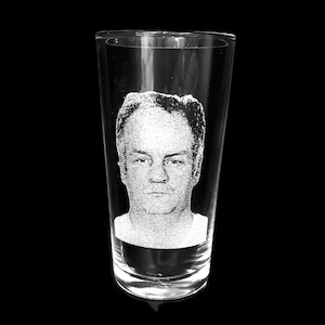 May include: A clear pint glass with a portrait etched into the glass. The portrait is of a man's face, and the glass is set against a black background.