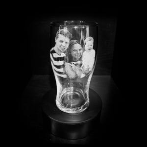 May include: A clear pint glass featuring an etched family portrait. The black and white image shows a man in a striped top, a woman, and a baby. The glass is displayed on a black, circular stand.