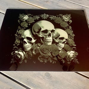 May include: Three white skulls with black eyes are surrounded by black and white roses on a black background. The skulls are arranged in a triangular shape with the largest skull at the top.