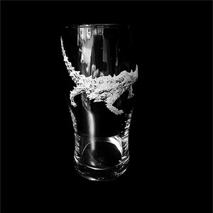 May include: A clear glass pint glass with a detailed etched design of an alligator. The alligator design is white and wraps around the glass. The glass is empty and set against a black background.