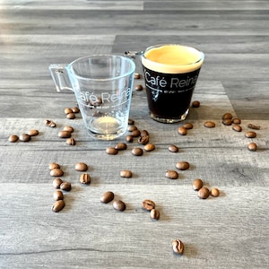 Può includere: Due tazze da espresso in vetro trasparente con la scritta "Cafe Reina est. 2020" su di esse. Una tazza è piena di caffè scuro e l'altra è vuota. I chicchi di caffè sono sparsi intorno alle tazze su una superficie di legno.
