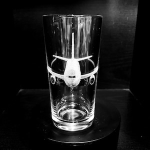 May include: A clear glass tumbler featuring an etched airplane design. The aircraft is shown from the front, with wings and engines. The cylindrical glass sits on a black base, ideal for serving drinks.