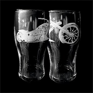 May include: Two clear glass beer mugs with etched motorcycle designs. One mug features a side view of a motorcycle, and the other displays a motorcycle wheel. The mugs are set against a black background.
