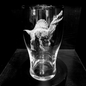 May include: A clear glass featuring an etched Spinosaurus dinosaur. The dinosaur is shown with its mouth open. The glass is set on a black base, creating a striking contrast.