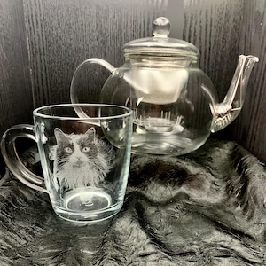 May include: A clear glass mug with a black and white cat etched into the side. The mug is sitting on a black fabric surface next to a clear glass teapot with a strainer.