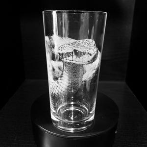 May include: A clear glass with a snake etched into the side. The glass is sitting on a black pedestal.