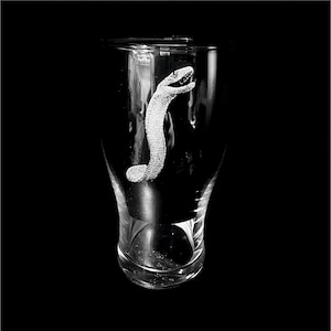 May include: A clear glass beer mug featuring an etched design of a snake. The snake is rendered in a textured style, with its head raised. The glass has a curved body and a thick base, set against a black backdrop.