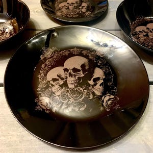 Könnte beinhalten: Set aus vier schwarzen Keramik-Dinnertellern mit Totenkopf- und Rosenmotiv. Die Teller haben eine glänzende Oberfläche und zeigen ein sich wiederholendes Muster aus drei Totenköpfen, die von Rosen umgeben sind.