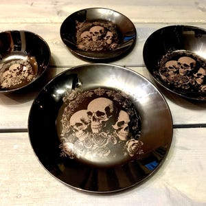 May include: Set of four black ceramic dinnerware featuring a skull and floral design. The plates and bowls have a glossy finish.