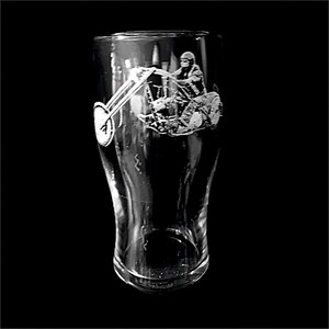 May include: A clear glass pint glass with a detailed etched design of a person riding a motorcycle. The motorcycle has a large front wheel and the rider is wearing a helmet. The glass is on a black background.