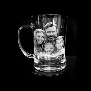 May include: A clear glass beer mug with a handle, featuring a family portrait etched onto the front. The black and white image shows two adults and two children. The mug is on a black surface.
