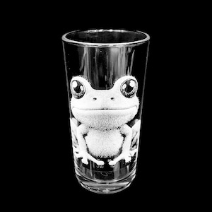 May include: A clear glass tumbler featuring a white etched graphic of a frog with large, round eyes and a wide, smiling mouth. The design is centered on the front of the cylindrical drinking vessel, which has a smooth rim and a solid base.