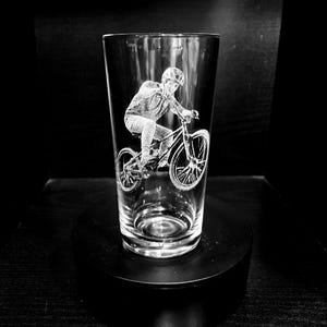 May include: Clear glass pint glass with an etched design of a person riding a mountain bike. The cyclist is wearing a helmet and riding on a bicycle with detailed wheels. The glass is displayed on a black surface.