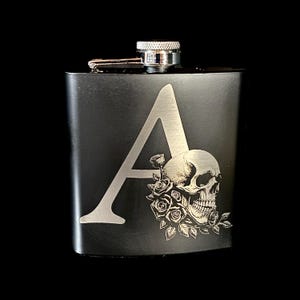 Puede incluir: Una petaca negra con una "A" plateada y un diseño de calavera con rosas. La petaca tiene un tapón y una bisagra plateados. El diseño está grabado en el metal. El fondo es negro.