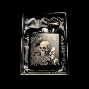 May include: A black stainless steel hip flask featuring a skull and rose design, presented in a black gift box with a satin lining. Includes a small funnel. The flask has a silver cap.