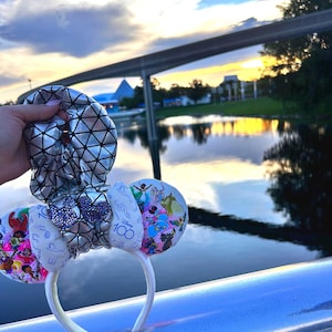 May include: A white headband with Mickey Mouse ears. The ears feature colorful Disney character designs and a silver bow. The background shows a sunset over a body of water and a monorail.