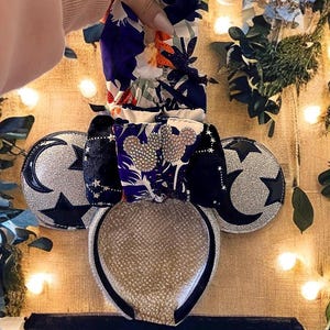May include: A pair of silver glitter Minnie Mouse ears with black velvet ears and a black headband. The ears are decorated with a silver crescent moon and a black star. The headband is covered in a black and white floral fabric.