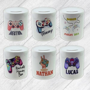 May include: Six white ceramic money boxes with different colourful designs featuring video game controllers and personalised names. The names on the money boxes are Hector, Tommy, Parker, Rachel's Money Box, Nathan, and Lucas.