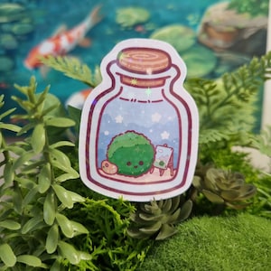 May include: A sticker of a glass jar with a green moss ball inside. The moss ball has a cute face and is holding a paintbrush. The jar is filled with a blue liquid and has white stars on it. The sticker is on a green background.