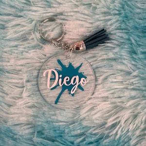 Op de afbeelding: Een sleutelhanger met een turquoise verfspatten en de naam "Diego" in witte letters. De sleutelhanger heeft een zilveren ring en een blauwe kwast.