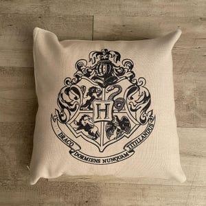 Può includere: Un cuscino beige con uno stemma nero di Hogwarts. Lo stemma presenta un leone, un tasso, un serpente e un'aquila. Il testo intorno allo stemma recita "Draco Dormiens Nunquam Titillandus".