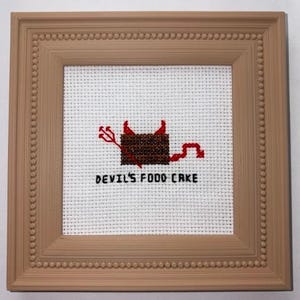 May include: A square cross-stitch embroidery in a light brown beaded frame. The white fabric features a brown rectangular cake with red devil horns, a red pitchfork, and a red tail. The black text below the cake reads DEVIL'S FOOD CAKE.