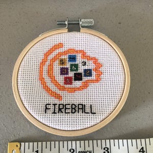 K&ouml;nnte beinhalten: Ein Kreuzstich-Stickrahmen mit dem Wort "FIREBALL" in Schwarz gestickt. Das Design zeigt eine orangefarbene Flamme mit bunten Quadraten. Der Holzrahmen hat einen Durchmesser von etwa 7,6 cm.