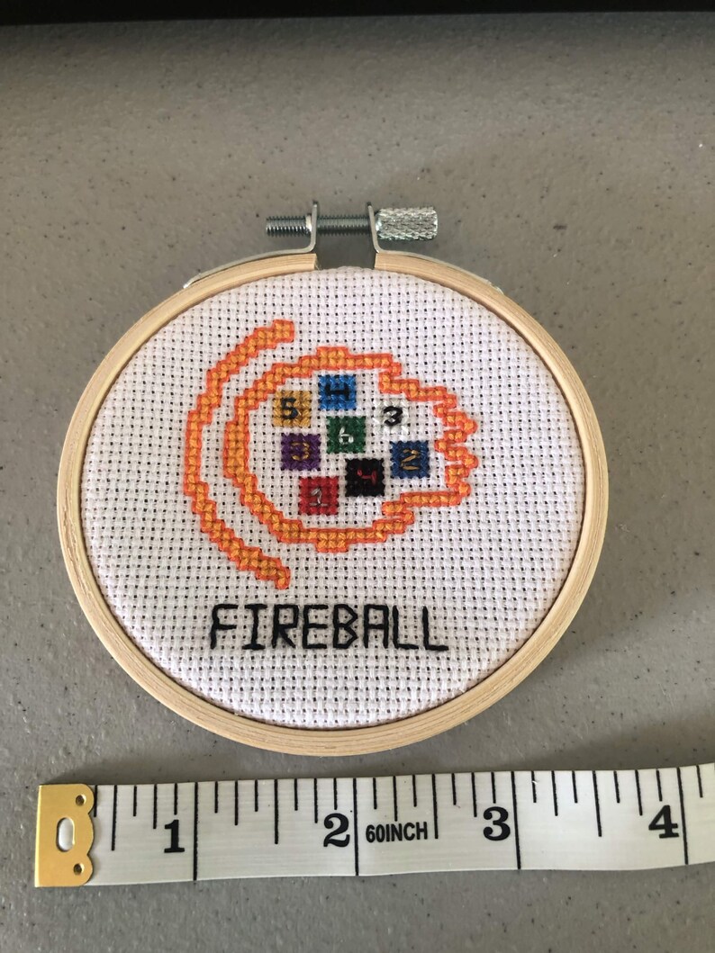 K&ouml;nnte beinhalten: Ein Kreuzstich-Stickrahmen mit dem Wort "FIREBALL" in Schwarz gestickt. Das Design zeigt eine orangefarbene Flamme mit bunten Quadraten. Der Holzrahmen hat einen Durchmesser von etwa 7,6 cm.