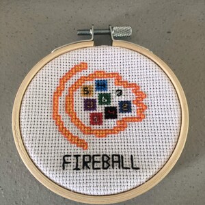 K&ouml;nnte beinhalten: Ein Kreuzstich-Stickrahmen mit dem Wort "FIREBALL" in Schwarz und einem orange-gelben Feuerball-Design. Das Design umfasst farbige Quadrate mit Zahlen. Die Stickerei befindet sich in einem Holzrahmen.