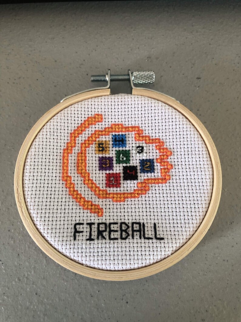K&ouml;nnte beinhalten: Ein Kreuzstich-Stickrahmen mit dem Wort "FIREBALL" in Schwarz und einem orange-gelben Feuerball-Design. Das Design umfasst farbige Quadrate mit Zahlen. Die Stickerei befindet sich in einem Holzrahmen.