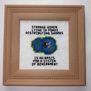 May include: A square cross-stitch embroidery in a light brown beaded frame. The white fabric features a blue pond graphic with a small figure and sword, surrounded by the black text: STRANGE WOMEN LYING IN PONDS DISTRIBUTING SWORDS IS NO BASIS FOR A SYSTEM OF GOVERNMENT.