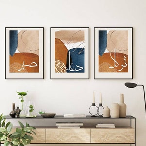 May include: Three abstract art prints with Arabic calligraphy in white on a brown, tan, and blue background. The prints are framed in black and hung on a white wall above a wooden console table.