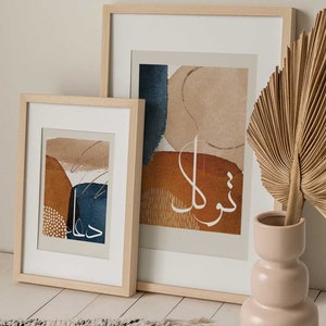 May include: Two framed abstract art prints with a beige background and brown, blue, and white abstract shapes. The prints are in wooden frames and are hanging on a white wall. A dried palm leaf is in a light pink vase on a white wooden floor.