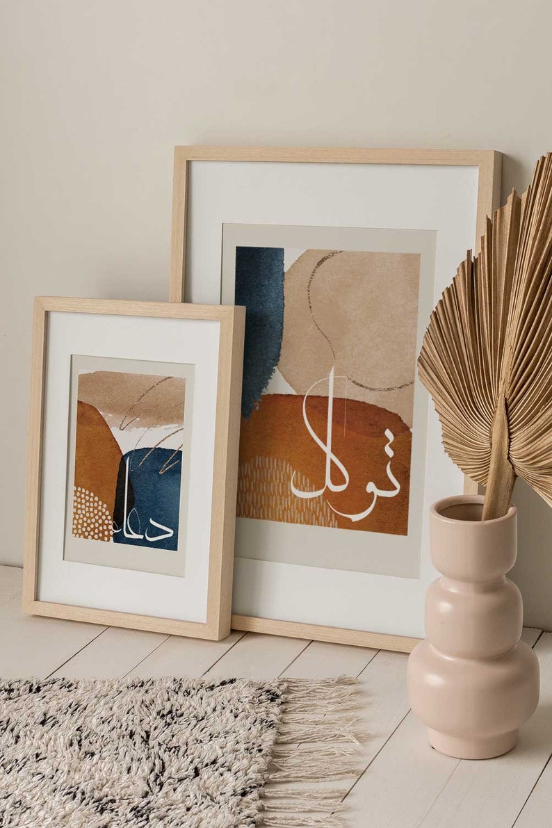 May include: Two framed abstract art prints with a beige background and brown, blue, and white abstract shapes. The prints are in wooden frames and are hanging on a white wall. A dried palm leaf is in a light pink vase on a white wooden floor.