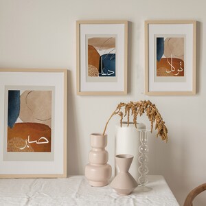 May include: Three framed abstract art prints featuring Arabic calligraphy and geometric shapes in shades of brown, beige, and blue. The prints are hanging on a white wall above a table with a vase of dried flowers.